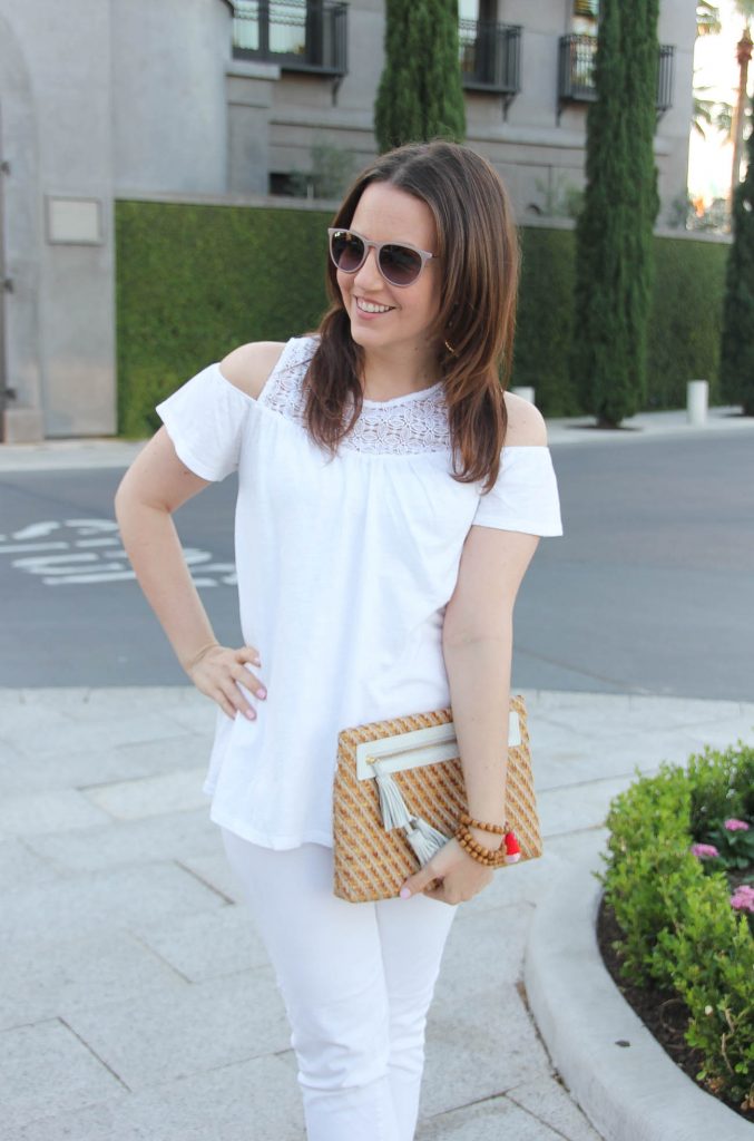 Lady in Violet, a Houston based style blogger, wears a spring white on white outfit including a cold shoulder top, white jeans, and a cute straw clutch with white tassels.